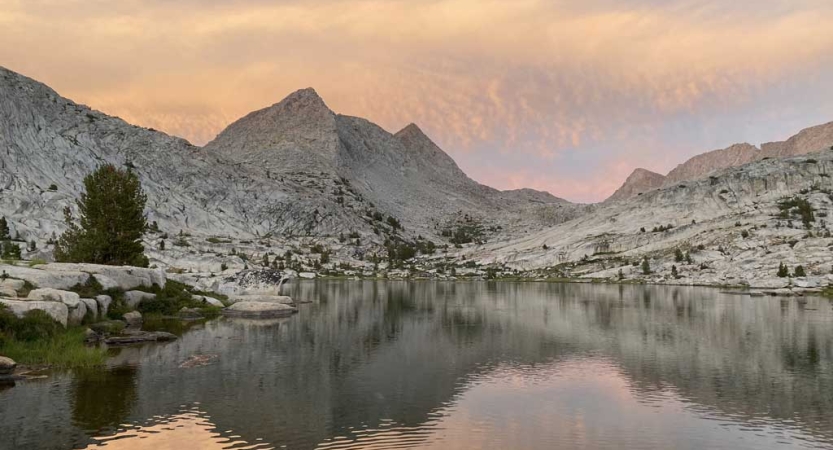 An alpine lake is nestled between mountains under a yellow, pink and purple sky.